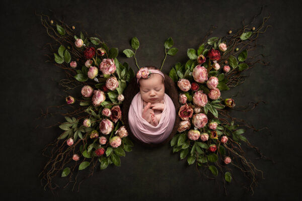 Newborn baby girl swaddled in soft pink wrap, posed in a Tampa photography studio on a white background with delicate flowers arranged in a butterfly shape around her.